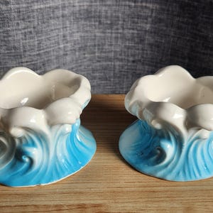 May include: Two ceramic egg cups featuring a sculpted wave design. The base of each cup is painted in a vibrant blue that transitions into a white, foam-like crest at the top. The interior of each cup is smooth, glossy, and finished in a solid white glaze.