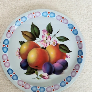May include: A round metal serving tray with a light blue background. The centre features a colourful illustration of peaches, plums, and pink blossoms with green leaves. The rim is decorated with a repeating pattern of blue and purple oval shapes.