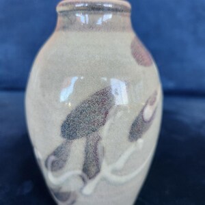 May include: A small, rounded ceramic vase with a smooth, glossy finish. The base colour is a muted, sandy beige, featuring abstract, dark purple-brown brushstroke patterns and thin, flowing white lines that wrap around the exterior surface.