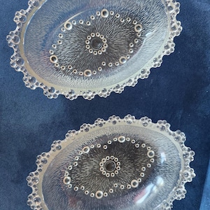 May include: Two oval clear glass dishes featuring a textured sunburst pattern in the centre, surrounded by a ring of small circular accents. The edges of each dish are decorated with a scalloped border of raised glass beads or bubbles.