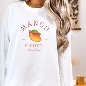 以下が含まれることがあります： マンゴーと「MANGO FESTIVAL Island Fresh」の赤いグラフィックと、マンゴーの下に「EST. 1985」の文字が書かれた白い長袖スウェットシャツ。