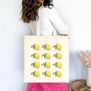 May include: A white canvas tote bag with a repeating pattern of yellow lemons and green leaves. The bag has two long handles.