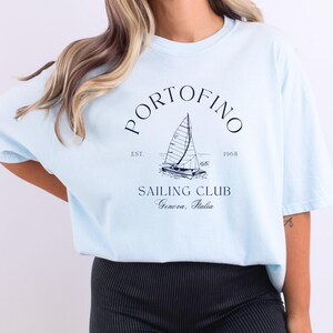 Peut inclure: Un t-shirt bleu clair avec un graphique de voilier et le texte "PORTOFINO SAILING CLUB EST. 1965 Genova, Italia".