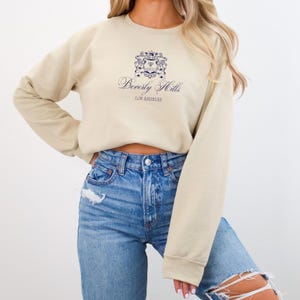May include: A beige cropped sweatshirt with a blue and white crest and the text "Beverly Hills, Los Angeles" printed on the front. The sweatshirt is being worn with blue jeans.