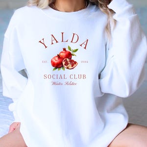 Puede incluir: Sudadera blanca con un diseño gráfico rojo que dice "Yalda Social Club" con una ilustración de granada y el texto "Est. 2024 Winter Solstice".