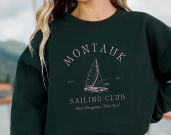 May include: A dark green sweatshirt with a white graphic design that says "Montauk Sailing Club" with a sailboat illustration and the text "Est. 1900 East Hampton, New York"