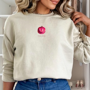 Puede incluir: Una sudadera de color verde claro con una ilustración de una granada roja y la palabra "YALDA" impresa en la parte delantera.