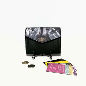 May include: Black leather wallet with a silver clasp. The wallet has a black and white patterned fabric flap. The wallet is sitting on a white surface with a few coins and some business cards.