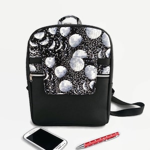 May include: Black backpack with a moon and stars pattern on the front pocket. The backpack has a zipped front pocket and a zipped main compartment. The backpack is sitting on a white surface with a mobile phone and a red pen.