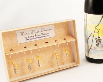 May include: A set of six wine glass charms in a box. The charms are silver with gold beads and a leaf design. The box is white with the text "Wine Glass Charms by Nancy Eden Designs www.nancyedendesigns.com". A bottle of wine and a wine glass with a charm are also shown.