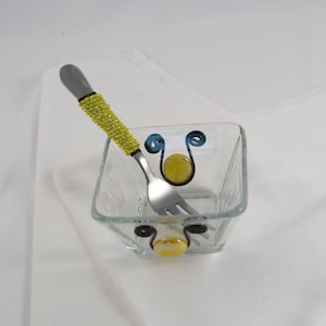 May include: A small square glass bowl with a blue and black design and two yellow glass accents. A fork with a yellow beaded handle is next to the bowl.