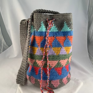 May include: A gray, handmade crochet bag with a colorful geometric pattern. The bag features a long strap and a drawstring closure with a braided cord. The pattern includes triangles in shades of blue, orange, pink, and green.