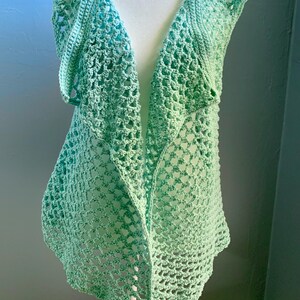 May include: A light green crochet vest with a wide, open neckline and a scalloped edge. The vest is made with a delicate, open stitch pattern.