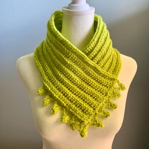 May include: A lime green crocheted cowl with a fringed edge. The cowl is folded over and draped around a white mannequin.