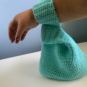 May include: A crocheted turquoise wrist cuff and matching bag. The cuff is worn on the wrist, and the bag is connected to the cuff. The crochet has a textured appearance. The items are a solid turquoise color.