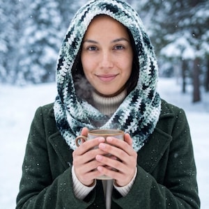 May include: A crochet hood in variegated shades of forest green, white, and black, worn over a beige turtleneck sweater and a dark green wool coat. The hood features a textured stitch pattern and is wrapped around the neck as a cowl.
