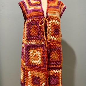 May include: A sleeveless, open-front crochet vest in a patchwork design. The vest features a mix of orange, purple, and cream-colored squares. A braided tie closure is at the waist. The garment is displayed on a mannequin.