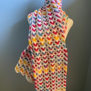 May include: A handmade, colorful crochet scarf displayed on a mannequin. The scarf features a repeating pattern of pink, blue, yellow, and white, with a scalloped edge. The scarf is made of soft yarn.