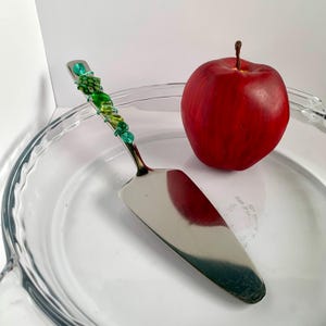 May include: A stainless steel pie server with a decorative handle wrapped in green wire and beads. A red apple sits next to the server on a clear glass pie dish. The handle features green beads and wire in a vine-like design.