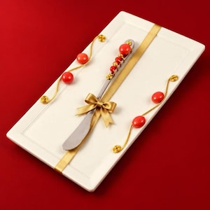 May include: A rectangular white serving tray featuring a silver spreader knife decorated with red beads and gold wire. A gold ribbon is wrapped around the tray, accented with a gold bow and additional red beads on gold wire swirls.