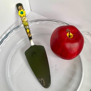 May include: A stainless steel pie server with a decorative handle featuring yellow and green beads and a yellow flower. A red apple sits next to the server on a clear glass pie plate.