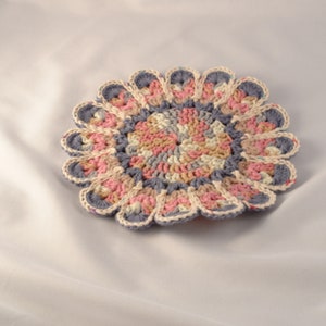 May include: A crocheted coaster with a circular design in shades of pink, beige, and blue. The coaster has a scalloped edge.