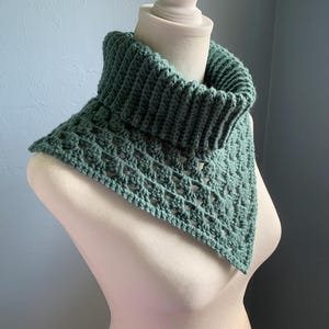 May include: A handmade, green crochet cowl scarf displayed on a mannequin. The cowl features a ribbed collar and an openwork design on the body. The scarf is a triangular shape and appears to be made of soft yarn.