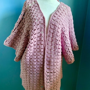 May include: A dusty rose crochet cardigan featuring an open front design, a V-neckline, and wide, loose-fitting sleeves. The garment is constructed with a repeating square stitch pattern throughout, creating a textured, lightweight, and airy appearance.