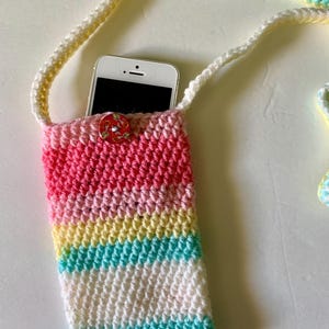 May include: A crocheted phone case with a pink, yellow, and blue striped design. The case has a white strap and a button closure. A smartphone is inside the case.
