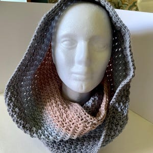 May include: A gray, pink, and white crocheted cowl with a hood. The cowl is made with a textured stitch and has a gradient of color from gray to pink.