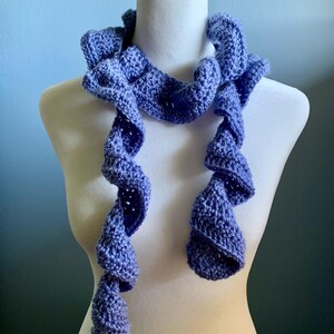 May include: A blue crocheted scarf with a ruffled design is displayed on a white mannequin. The scarf has a spiral shape with long, dangling ends. The color is a mix of light and dark blue, creating a textured appearance. The scarf is a handmade accessory.