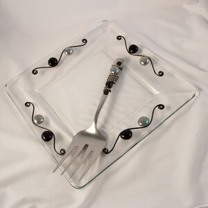 Puede incluir: Un plato cuadrado de vidrio transparente con un tenedor de cuentas negras y plateadas. El plato tiene un diseño decorativo de alambre negro con cuentas negras y plateadas.