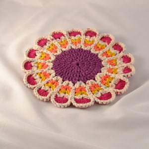 May include: A crocheted flower-shaped coaster in shades of pink, orange, yellow, and purple. The coaster has a raised center and a scalloped edge.