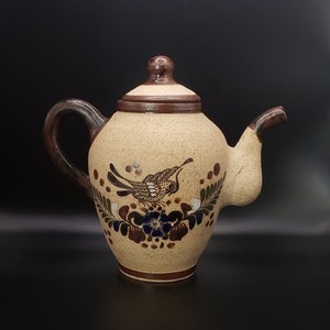 Pode incluir: Uma chaleira de cerâmica marrom com um design de pássaro e flores. A chaleira tem uma alça e um bico marrons e um corpo bege com detalhes pintados em marrom, verde, azul e branco.