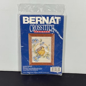 Pode incluir: Um kit de ponto cruz com um pássaro chamado 'Canada Warbler' com flores azuis em uma moldura circular. O kit inclui tecido Aida de 14 pontos, agulha, instruções e fio suficiente para completar o desenho. O kit é da Bernat e está etiquetado como "95-4241-00".