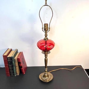 May include: A table lamp with a brass base and stem featuring a central red glass globe with etched floral patterns. The lamp includes a brass socket and a metal harp for a shade. The lamp is positioned next to a small stack of four vintage books.