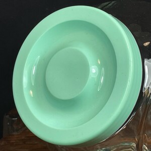 May include: A light green, circular lid with a recessed center. The lid has a smooth, glossy finish and appears to be made of plastic or ceramic. The lid is likely for a jar or container, and is shown against a dark background.