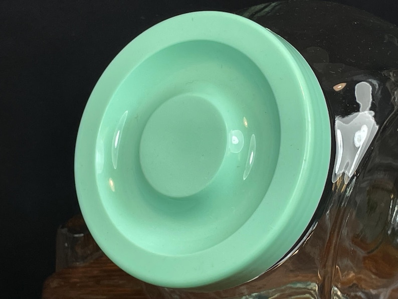 May include: A light green, circular lid with a recessed center. The lid has a smooth, glossy finish and appears to be made of plastic or ceramic. The lid is likely for a jar or container, and is shown against a dark background.
