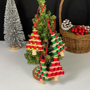 Puede incluir: Tres adornos navideños hechos a mano con carretes de hilo rojo y verde. Los adornos están decorados con detalles rojos y verdes y cuelgan de cintas rojas. Un pequeño árbol de Navidad y una cesta con piñas y bayas rojas están en el fondo.
