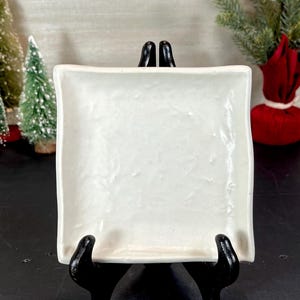 May include: A square, off-white ceramic dish with slightly uneven edges. The dish is displayed on a black stand, with miniature Christmas trees and festive decorations in the background.