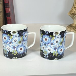 May include: Two ceramic mugs with white handles and interiors. The exterior of each mug is black and covered in a floral pattern of blue, white, and yellow flowers. The mugs are sitting on a white surface.