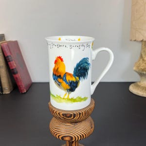 Puede incluir: Taza de cerámica blanca con una ilustración de gallo colorido. El gallo tiene una cresta roja, cuerpo amarillo y naranja y plumas de cola azules. La taza tiene un asa curva y el texto "My feathers, Scratchy toes".