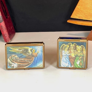 May include: Two vintage brass boxes with hinged lids. One box features an angel illustration with a flowing robe and a bright light. The other box displays a group of angels in colourful robes. Both boxes have a rectangular shape.
