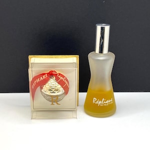 May include: A vintage perfume bottle with a silver cap and a frosted glass body, next to a small box with a gold trim and a red ribbon. The perfume bottle has the word "Réplique" printed on it.