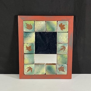 Può includere: Uno specchio con cornice rossa con un mosaico di piastrelle verdi e gialle con motivi di foglie marroni.