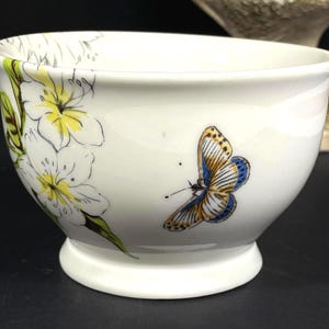 Op de afbeelding: Een witte keramische kom met een bloemen- en vlinderontwerp. De kom heeft een brede rand en een afgeronde basis. De bloemen zijn geel en wit met groene bladeren. De vlinder is blauw en geel met bruine markeringen.