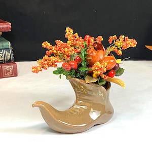 Könnte beinhalten: Eine braune Keramikvase in Stiefelform, gefüllt mit bunten Kunstbeeren und Blättern. Die Anordnung umfasst orange, gelbe und rote Beeren mit grünen Blättern. Die Vase hat einen warmen, erdigen Farbton.