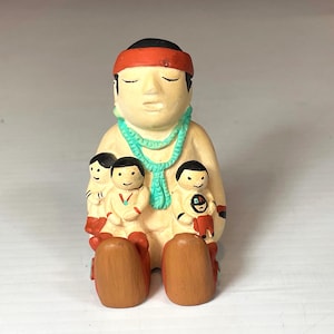 May include: A ceramic figurine of a Native American person with a red headband and turquoise necklace, holding three smaller figurines of children.