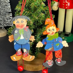 May include: Two wooden Christmas tree decorations. One features a cartoon figure with black hair, a blue jacket, and clogs. The other has red hair, a blue coat, and orange trousers. Both have red hanging strings and red ball feet.