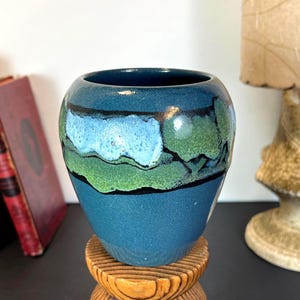 Könnte beinhalten: Eine blaue Keramikvase mit runder Form. Die Vase hat ein gemaltes Design mit blauen, grünen und schwarzen Akzenten. Die Vase steht auf einem Holzständer. Dekorative Wohnkultur.